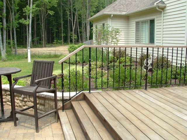 Custom iron rail at backside of deck leading to brick patio with stone wall and landscape plantings in the backround.